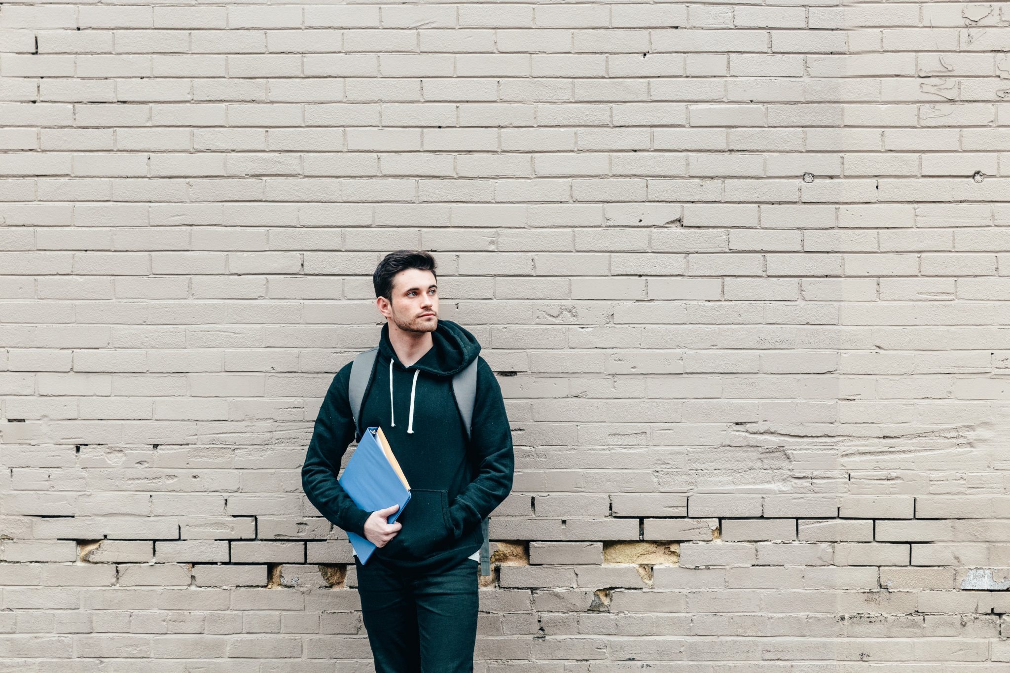 Male student leaning against a brick wall holding textbooks