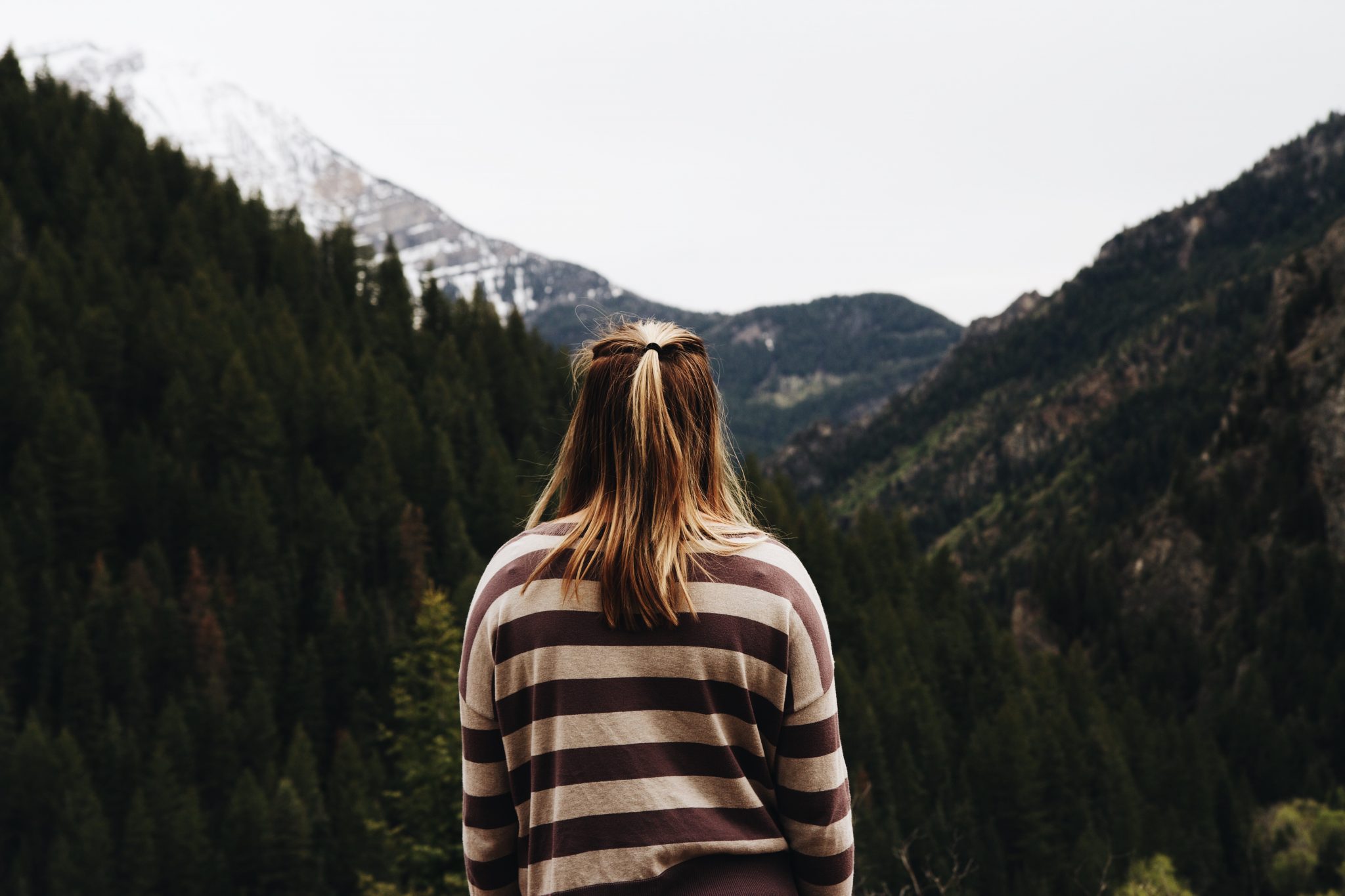 A blonde woman stands with her back facing the camera looking out towards a forested, mountain range.